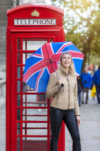 Portrait of smiling woman holding umbrella while standing on footpath in city