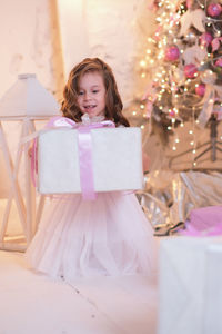 A beatiful girl with curly hair in white dress is surprising and holding christmas box gift 