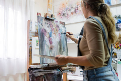 Side view of crop female artist standing near easel and drawing colorful picture with paints while working in creative workshop