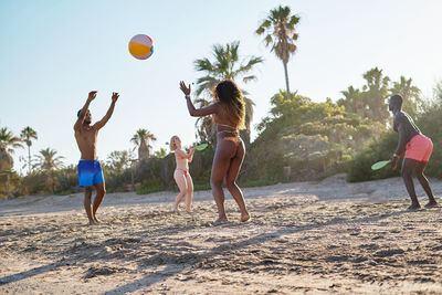 Low angle of diverse men and women tossing ball and playing beach tennis while spending summer weekend day on sandy seashore together