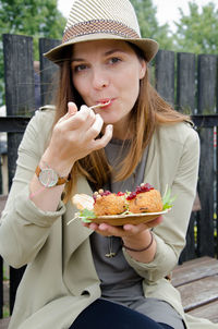 Portrait of beautiful woman eating salad while sitting on bench against fence