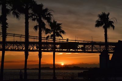 Silhouette bridge over sea against dramatic sky during sunset