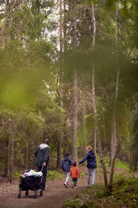 Rear view of man pulling camping cart while hiking with family amidst trees in forest