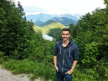 Portrait of smiling young man with hands in pockets standing on mountain in forest