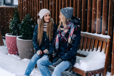 Two women friends in winter coats and hats sitting on a bench on the terrace look at each other.