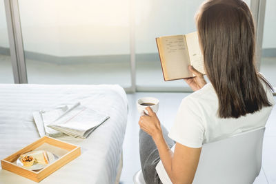 Rear view of woman having coffee while reading book at home