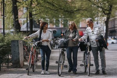 Male and female senior friends talking while wheeling bicycles on footpath