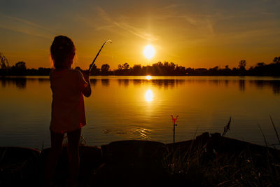 Silhouette girl fishing at lake against sky during sunset