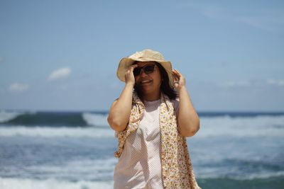 Young woman wearing hat standing at beach against sky