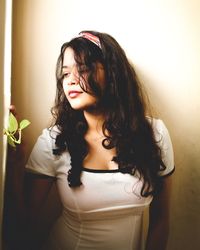 Young woman looking away while standing against wall at home