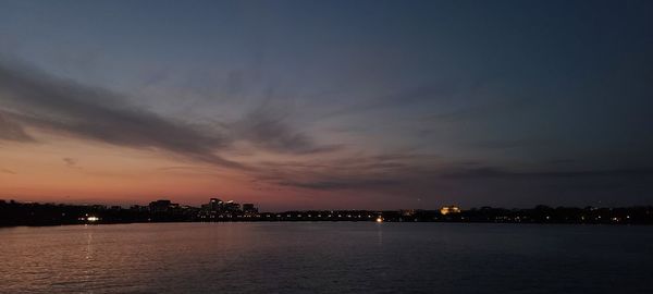 Illuminated city by sea against sky at sunset