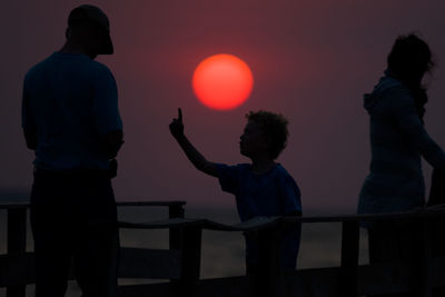 Silhouette boy talking to man while standing on pier against sky during sunset