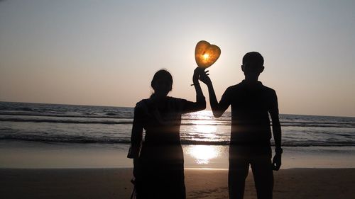 Silhouette couple with heart shaped balloon standing at beach during sunset