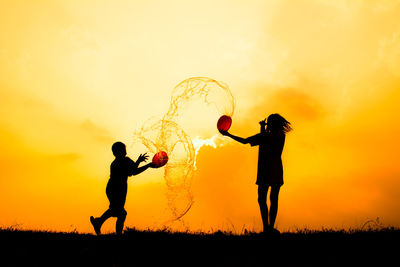 Low angle view of silhouette siblings playing with water against orange sky