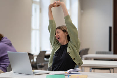 Tired female mature student taking break stretching arms while studying online in university library