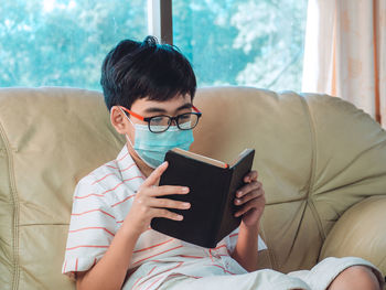 Boy wearing mask reading book sitting on sofa at home