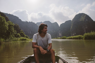 Man looking away while sitting in boat on river against sky