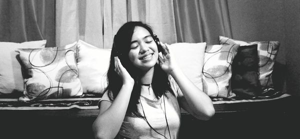 Smiling young woman listening music through headphones against sofa at home