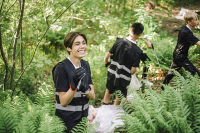 Cheerful teenage boy wearing gloves standing amidst green plants