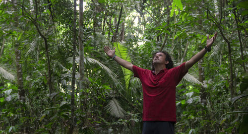 Young tourist man raises his arms and looks up to the sky in the middle of an amazonian forest