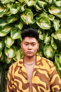 Dreamy thoughtful young filipino non binary man wearing cool shirt with earring standing in green garden looking at camera