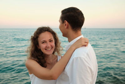 Young adult woman embraces her beloved man smiling against seaside sunset and looking to a camera