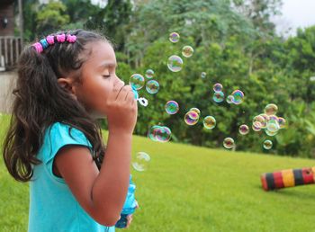 Side view of girl blowing bubbles on grassy field against trees