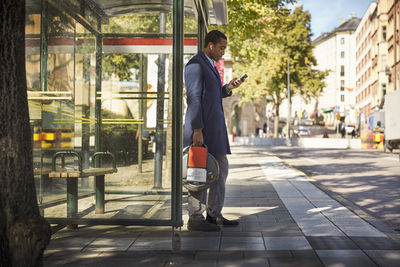 Full length side view of young male commuter using smart phone while holding electric unicycle at bus stop in city