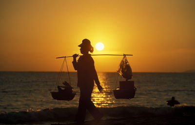 Silhouette women vendor walking at beach against sky during sunset