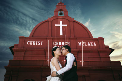 Bride and groom embracing in front of church against sky