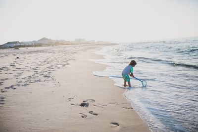 Side view of boy playing with fishing net in sea against clear sky during sunset