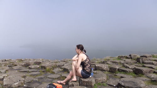 Woman sitting on rock against sky during foggy weather