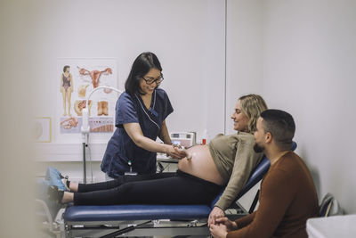 Smiling gynecologist doing ultrasound of pregnant woman sitting by man at hospital