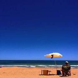 Rear view of man sitting on chair at beach against clear blue sky