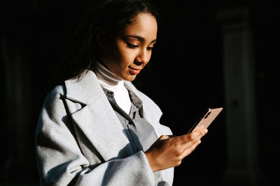 Young ethnic female in stylish casual outfit messaging on mobile phone while standing on urban street in sunny autumn day