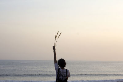 Rear view of a young woman holding flower plant with arm raised on beach against sky during sunset