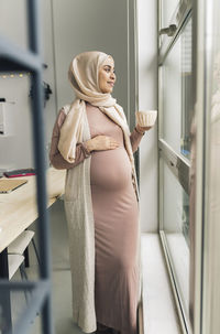 Thoughtful pregnant woman looking through window at home