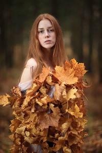 Portrait of young woman covered with leaves standing at forest during autumn