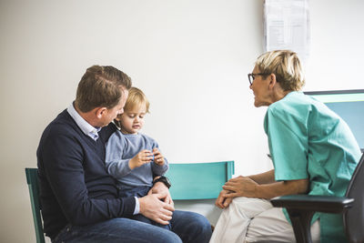 Side view of mature female doctor talking to boy with father in clinic