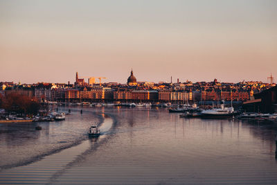 Boats in river by buildings against sky during sunset