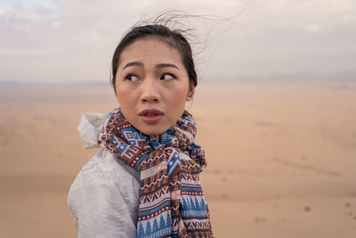 Side view of pensive young ethnic female tourist contemplating natural landscape while sitting on sandy dune in desert in saudi arabia looking away