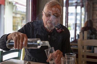 Senior man smoking cigar pouring drink in glass sitting at table
