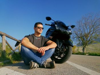 Man sitting by motorcycle on road against clear blue sky