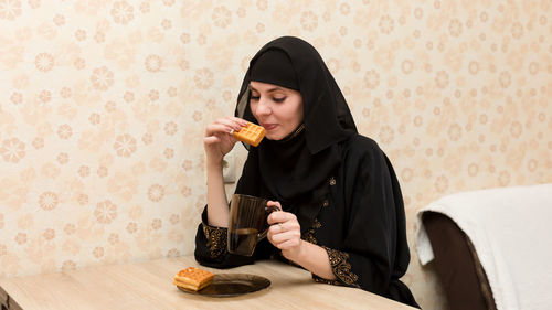 Muslim woman in national clothes has breakfast with tea and cookies at kitchen table.
