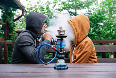 Man and woman sitting on table by tree and smoking hookah vipe