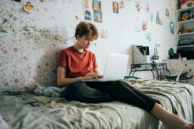 Teenage girl sitting on bed studying online using laptop.