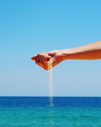 Cropped hand of person holding sand over sea against clear sky