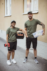 Full length portrait of smiling male and female workers with toolboxes by building