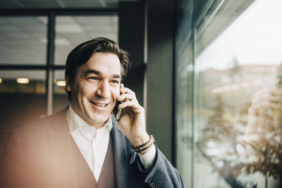 Mid adult businessman talking on smart phone while standing by window in office