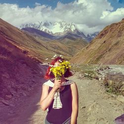 Woman hiding face with flowers while standing against mountains